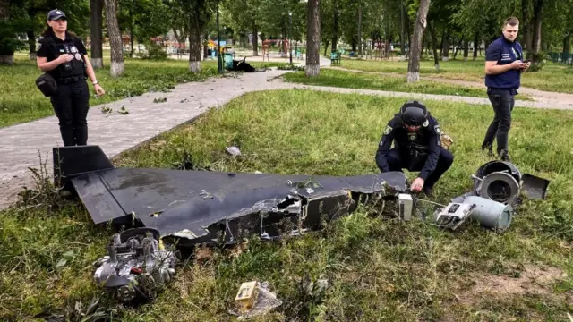 黒っぽい制服を着たウクライナの当局者数人が、歩道近くの草の上に残された黒っぽいドローンの残骸を調べている