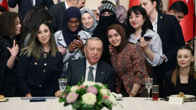 Cumhurbaşkanı Erdoğan masada oturuyor, etrafında genç kadınlar gülümsüyor