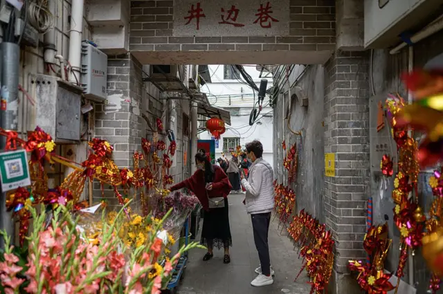 Люди идут вдоль стен, украшенных гирляндами и цветами