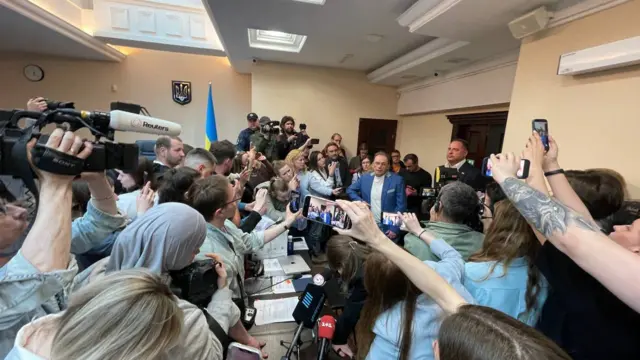 Велике світле приміщення, заповнене людьми. У центрі — довгий стіл, за яким сидять і стоять кілька осіб у діловому одязі. Навколо щільно зібрані журналісти з камерами, мікрофонами та смартфонами, багато рук підняті для зйомки. Зліва видно професійну відеокамеру з логотипом Reuters, на столі – мікрофони телеканалів "Прямий" і 1+1.