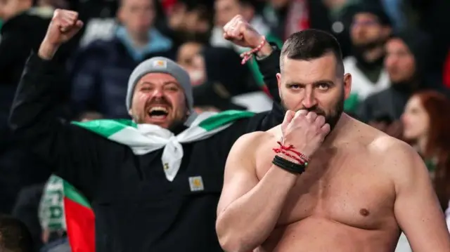 مشجعان لكرة القدم في مدرج على اليمين شاب بلحية دون قميص يرفع قبضته إلى فمه، على اليسار شاب يضع علم فريق حول رقبته، برتدي قبعة ويرفه يديه محتفلاً