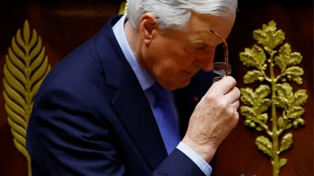 French Prime Minister Michel Barnier remove im glasses. E dey wear a navy suit and a patterned light blue tie and shirt.