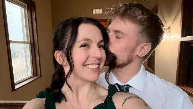 Ashley tem cabelo castanho escuro e está tirando uma selfie com Joe, que beija o rosto dela. 