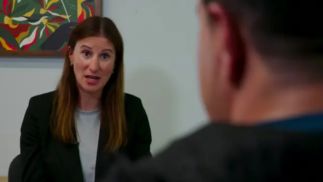 Reporter Helen Drew wearing a dark blazer speaks during an interview, filmed over the shoulder of the person opposite her