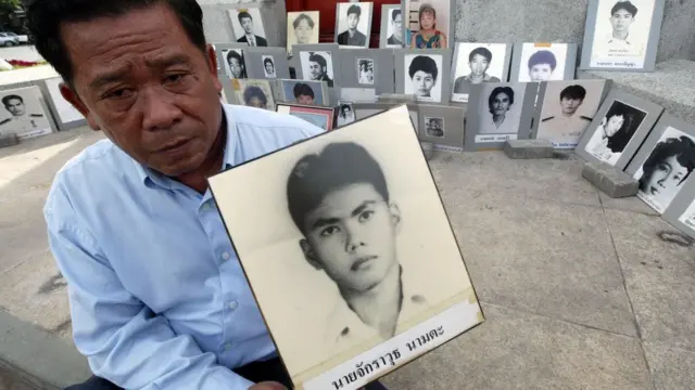 พ่อผู้สูญเสียลูกชายในช่วงพฤษภาทมิฬ ถือรูปลูกชายของเขาเบื้องหลังคือรูปของผู้ที่เสียชีวิตและสูญหายจากเหตุการณ์เดียวกัน