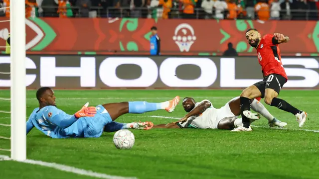 Mohamed Salah marque un but pour l'Égypte contre la Côte d'Ivoire lors de la CAN 2025. Salah, vêtu d'un maillot rouge de l'Égypte, d'un short et de chaussettes noirs et de crampons blancs, se trouve à l'extrême droite de l'image, tombant légèrement sur sa gauche tout en regardant le ballon se diriger vers le but. Un défenseur ivoirien tout de blanc vêtu glisse sur le sol dans une tentative infructueuse de bloquer le tir, tandis que le gardien Yahia Fofana, vêtu de bleu ciel, se trouve également sur la pelouse verte. Le ballon traverse le terrain, passe devant les trois joueurs et se dirige vers le but, que l'on aperçoit partiellement à l'extrême gauche de l'image.
