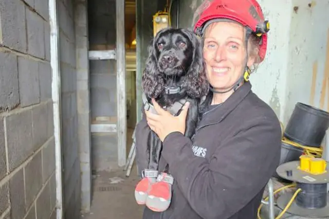 Una mujer con un casco rojo y una chaqueta negra con las letras WMFS sostiene un cocker spaniel negro que lleva botas de protección rojas y grises.