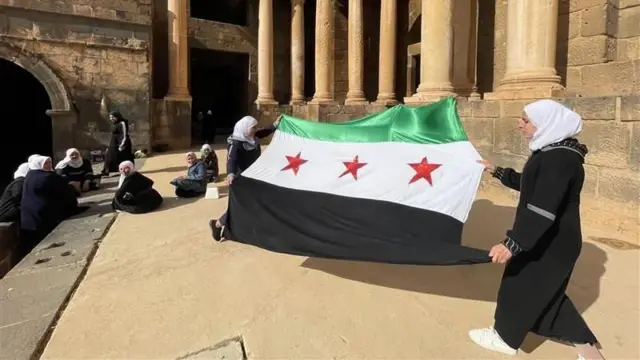 Las mujeres observan cómo se despliega la bandera siria utilizada por la oposición con una franja verde, una franja blanca con tres estrellas rojas y una franja negra.