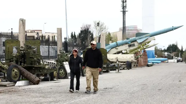 люди гуляют по парку, где стоят ракеты и другая военная техника