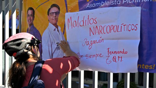 Una mujer pega un aviso con el mensaje Malditos Narcopolíticos pagarán sobre la imagen de Fernando Villavicencio 