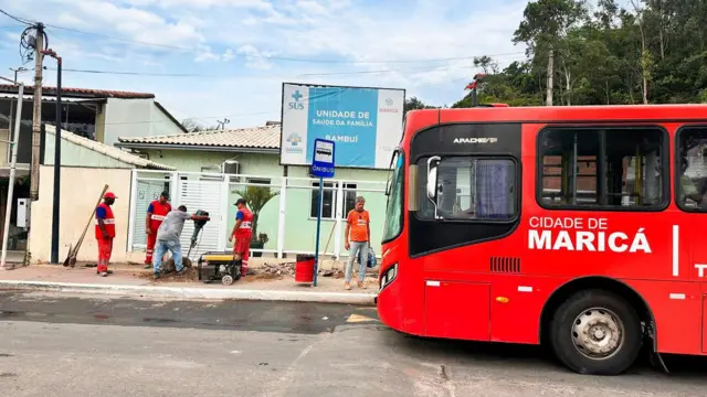 Ônibus vermelho gratuito da cidade de Maricá, em frente a uma Unidade de Saúde da Família, com operários vestidos em uniformes laranjas trabalhando em obra na calçada