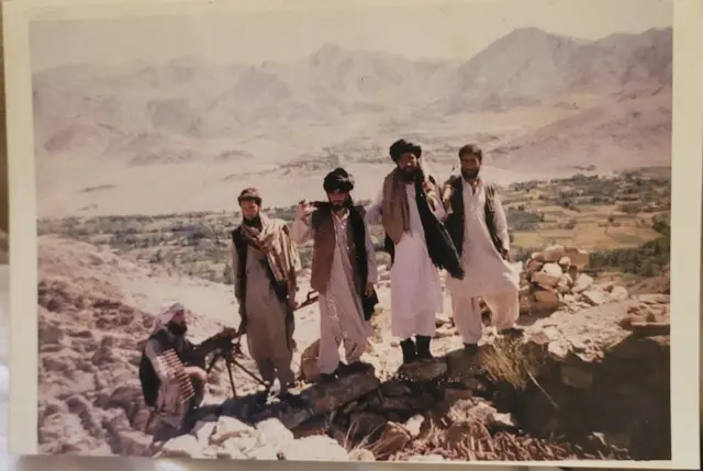 میوند بنايي، په ښۍ خوا کې (بې لونګۍ کس چې کلاشینکوف ور سره دی) د طالبانو له غړو سره. ۱۳۷۶ کال، میدان ښار، د خروټو کلی 