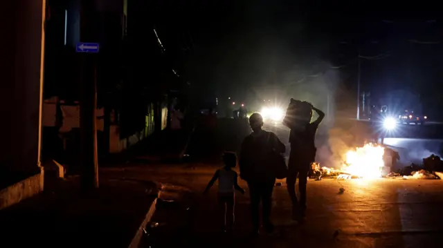 Havana’da bir yolun ortasında yanan çöp yığını önünde duran üç çocuk; sokak, uzaktaki araç farları dışında tamamen karanlık