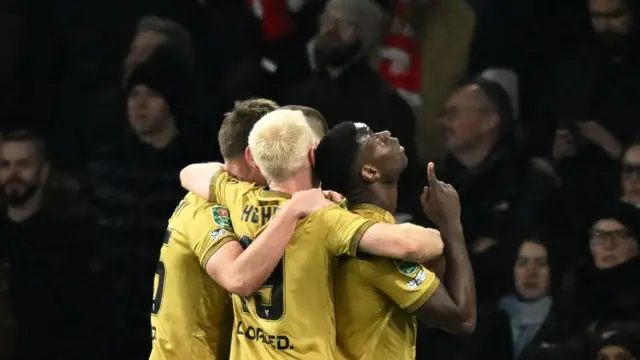 Crystal Palace Marc Guehi dey look up to di sky and dey point. Several of im team mates dey hug am from behind. Dem dey all wear gold jerseys. 