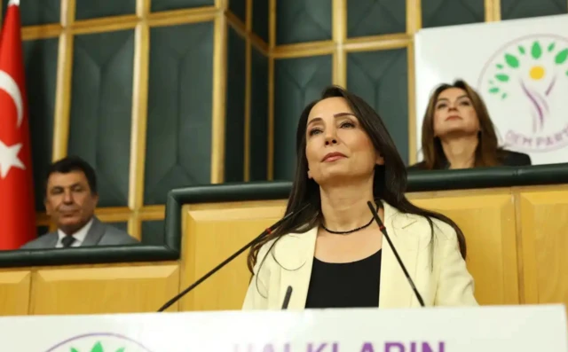 DEM Parti grup toplantısında bir kadın siyasetçi beyaz takımıyla kürsüde, arkada bir uzun saçlı siyasetçi DEM logosu altında oturuyor.