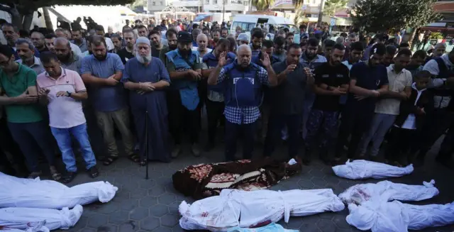 Wael Al-Dahdouh leads prayers at his family's funeral with dozens of people behind him as he raises his hands above bodies wrapped in shrouds
