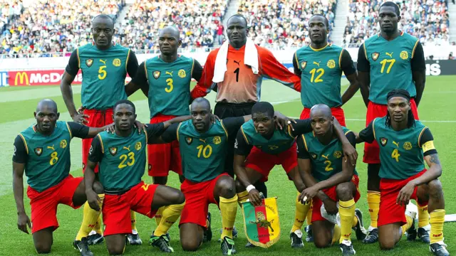 Photo de l'équipe du Cameroun avant un match de la Coupe du monde 2002. Les joueurs de champ extérieur portent une tenue composée de chaussettes jaunes, d'un short rouge et d'un maillot vert sans manches sur lequel sont cousues des manches noires sur le haut des bras. Le gardien de but porte un haut orange et un short noir