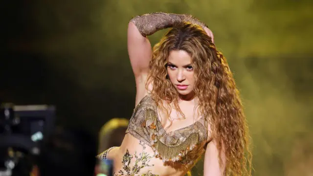 Shakira se apresenta no palco do Grammy com expressão intensa e olhar fixo, usando um figurino dourado brilhante que destaca o movimento do corpo; seus cabelos longos e ondulados caem sobre os ombros enquanto ela posa com um braço levantado sobre a cabeça, em meio a uma iluminação quente.