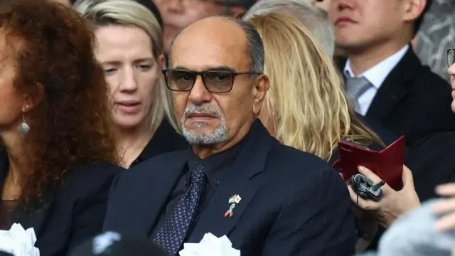 ダークスーツに白い胸花をつけたパレスチナ代表部のシアム代表。胸に日本とパレスチナの旗のピンバッジをつけている