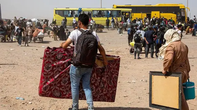 یک مرد بازگشتی افغان با پیراهن سفید و پتلون آبی که یک بیگ سیاه در پشت دارد و وسایل خانه اش را در دست دارد و یک زن با چادر روشن و لباس قهوه‌ای در یک دست یک قاب عکس بزرگ و در دست دیگر ترموز آب دارد در میان جمعیت بازگشتی به افغانستان 