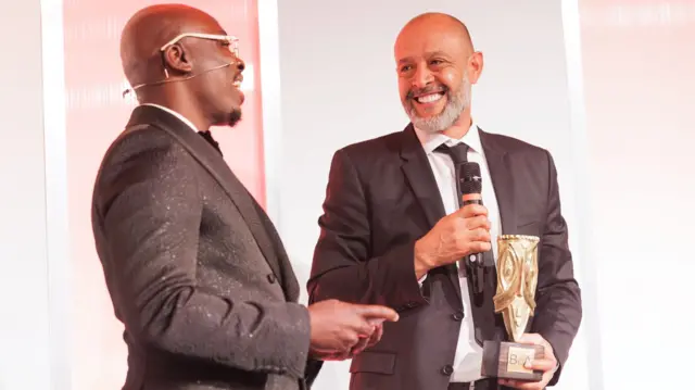 Edi Kadi et Nuno Espirito Santo, vêtus de costumes, apparaissent en plan serré, de la taille jusqu'à la tête, lors du dîner de gala des Best of Africa Awards 2025. Kadi, qui porte également des lunettes à monture blanche, prend la parole tandis que Nuno, souriant, tient un micro noir dans sa main droite et le trophée doré Best of Africa dans sa main gauche. Sur scène, les deux hommes tournent légèrement la tête pour se regarder.