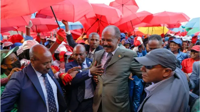 Perezida wa Eritrea Isaias Afwerki akikijwe n'abantu bafite imitaka y'ibara ritukura. Arimo kumwenyura