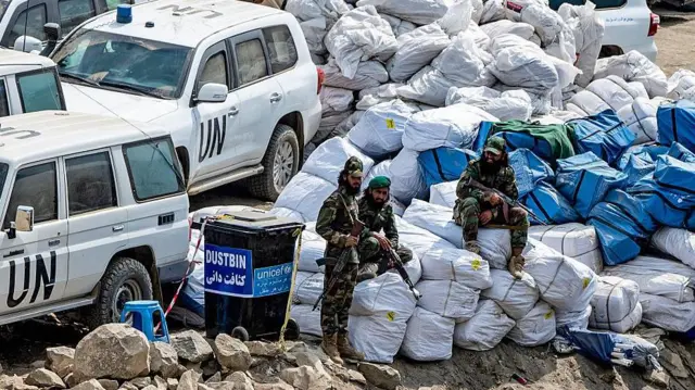 کمک‌های ملل متحد در خریطه‌های سفید و آبی که دو سرباز طالبان سلاح به‌دست بالای آن نشسته و یکی دیگر سلاح به‌دستش در کنار آن ایستاده است. آن طرف چند موتر ملل متحد پارک اند.