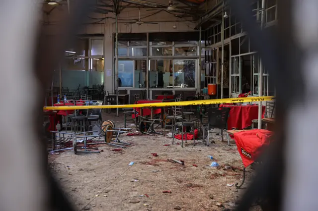 Yuvarlak bir deliğin içinden patlama yaşanan kafe, yere dağılmış sandalyeler ve yerde kan izleri görülüyor. Önüne sarı polis kordonu çekilmiş.