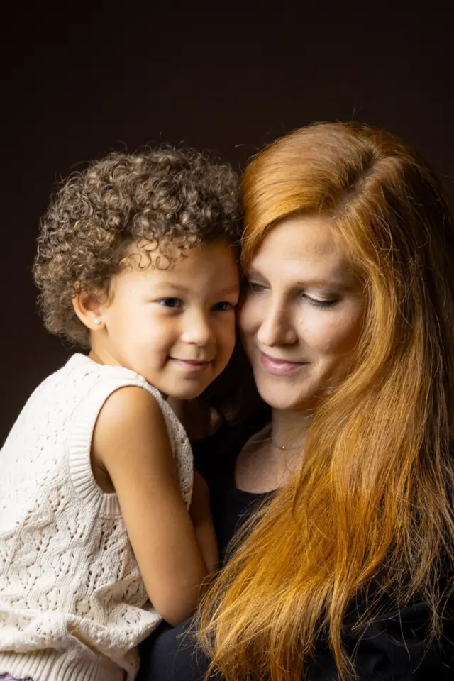 Retrato de estudio en primer plano de una mujer blanca con cabello largo y rojo que sostiene a una niña pequeña de piel morena y con cabello corto y rizado. 