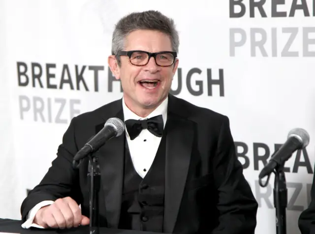 Andrew Strominger, con lentes, vestido de negro, con una camisa blanca y un corbatín negro, está sentado, en el fondo se lee en letras grandes: Breakthrough Prize