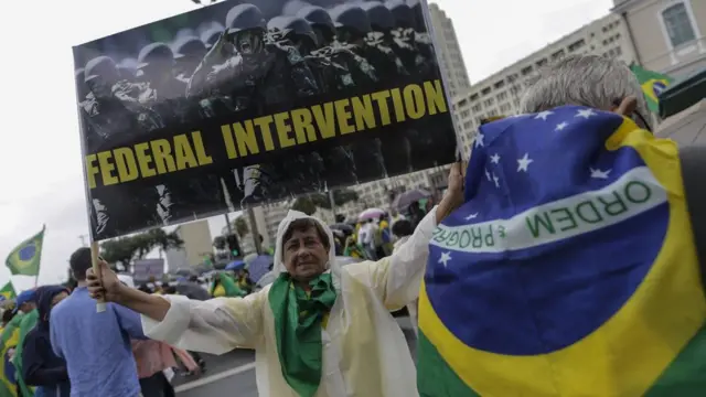 Senhora na rua levanta cartaz com fotocomo fazer aposta no jogo de futebolmilitares e pedidocomo fazer aposta no jogo de futebolinglês por 'Federal intervention'