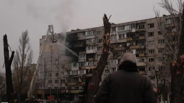 Пошкоджений обстрілами будинок навпроти якого стоїть людина спиною до камери. Обпалені вогнем дерева 