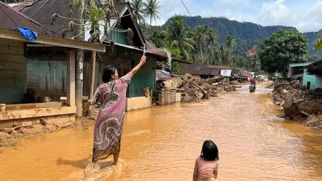 Seorang perempuan menghadap ke belakang sambil membuat gelembung busa sabun, dan diperhatikan oleh anak kecil di belakangnya di tengah jalan yang tergenang air.