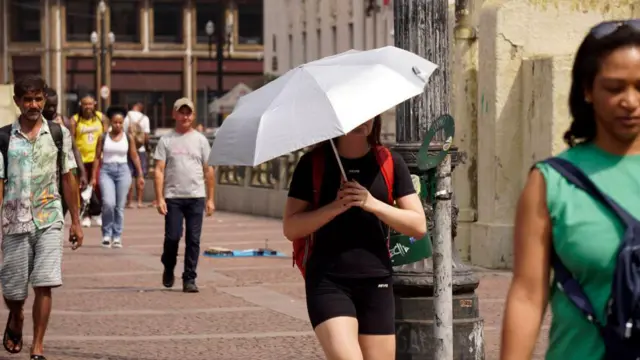 Pessoas caminham na rua durante onda de calor em São Paulo. Uma delas usa um guarda-chuva para se proteger do sol