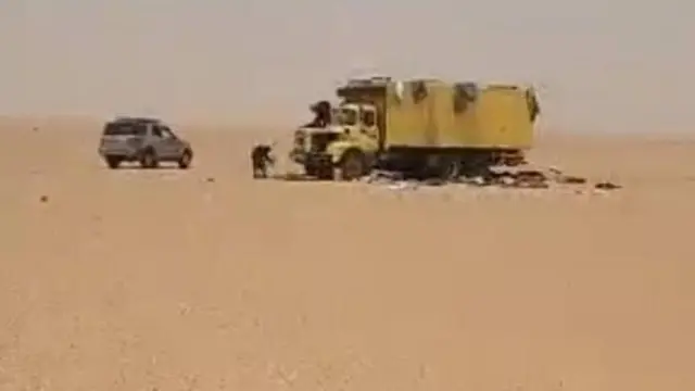 Un camion jaune et un SUV gris sont garés dans un vaste paysage désertique, avec quelques personnes debout à proximité sur le sable