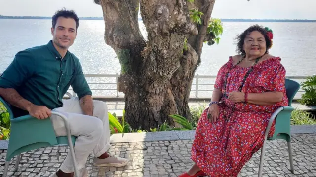 Dona Onete com vestido vermelho estampado se senta à frente do repórter João Fellet, que veste camisa verde e calça clara; ao fundo deles, vê-se o tronco de uma árvore e um rio bastante largo