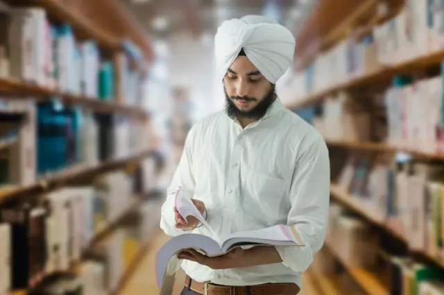 ਚਿੱਟੀ ਪੱਗ ਅਤੇ ਕਮੀਜ਼ ਪਹਿਨੇ ਲੜਕਾ, ਲਾਇਬ੍ਰੇਰੀ ਵਿੱਚ ਖੜ੍ਹਾ ਕਿਤਾਬ ਫਰੋਲ ਰਿਹਾ ਹੈ