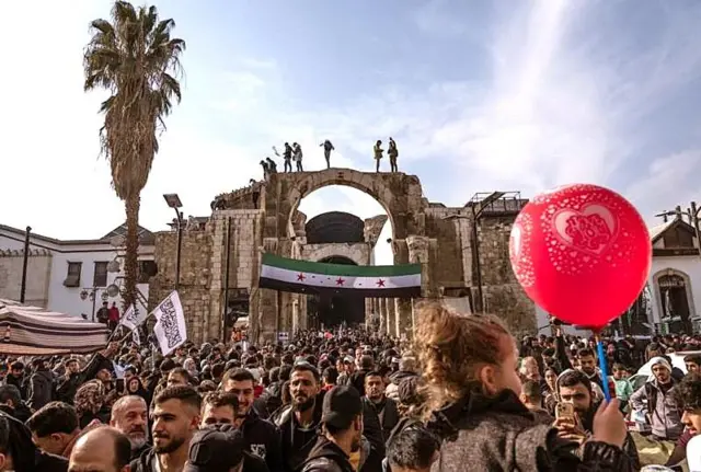 Suriye bayrağı önünde kalabalıklar, kırmızı balon tutan bir çocuk, palmiye ağacı