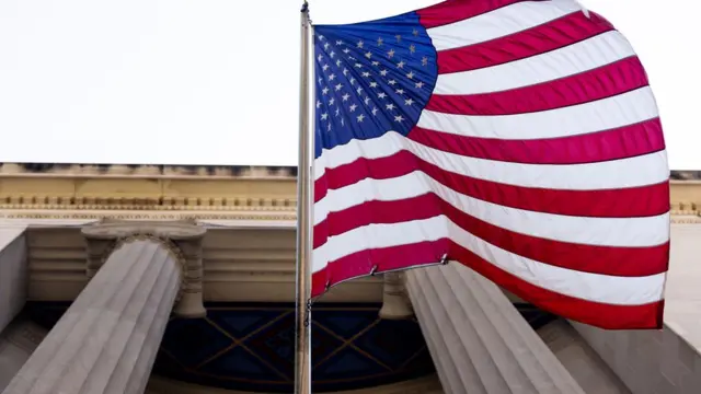 Fotografia colorida mostra bandeira dos EUA fotografada por baixo na entradas de um prédio com colunas gregas