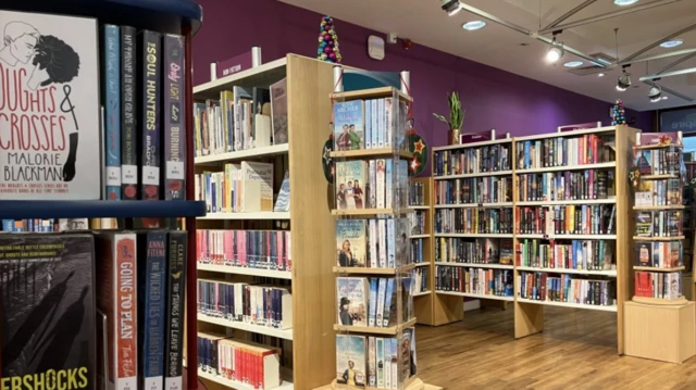 Varias estanterías de madera están llenas de libros. Las paredes están pintadas de morado y el suelo es de madera.
