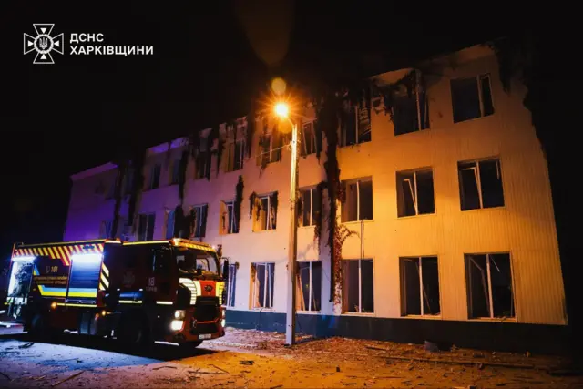 Біла триповерхова будівля з вибитими вікнами. біля неї стоїть пожежна машина