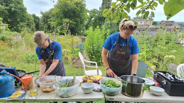 Dos personas cocinan en dos mesas cubiertas de comida recolectada. Están en un jardín verde en lo que parece un día soleado.
