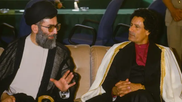 Gaddafi , sitting to the right of the image, looks at Khamenei as the Iranian leader talks. Gaddafi is holding his hands together on his lap. He is wearing a white robe with golden embroidery on the sides, a black jacket and dark red shirt with no collar, buttoned to the top. Khamenei is holding his hand up as he speaks. He is wearing black clothing on his head, a grey and black jacket and cream shirt. The pair are sitting next to each other on a sofa, with desk chairs and a meeting table behind them.