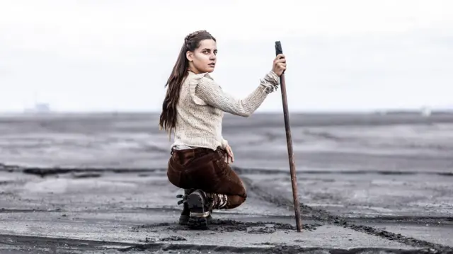 Девојка у чучњу у смеђим панталонама и белој мајици окружена пустињом држи штап у руци и гледа у камеру - Воља синовљева