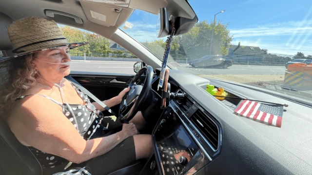 Marta Helena usando chapéu, dentro do seu carro, com uma bandeira dos Estados Unidos