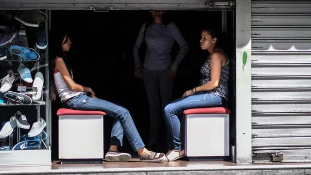 Funcionárias de uma sapataria sentadas na entrada de uma loja às escuras, durante um corte de eletricidade em Caracas