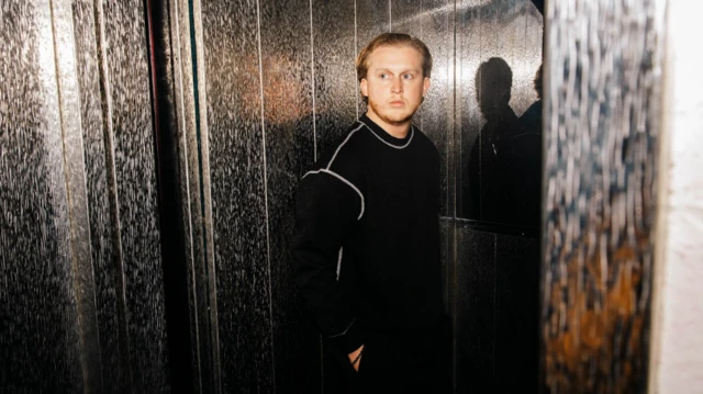 A professional, and stylised, photo of Max Dean, the musician, standing in front of a dark wall with a marbled-stripe design. He is looking away to the left of the shot and is wearing a black jumper