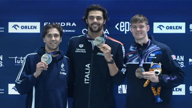 Tres hombres, todos con chándales oscuros de sus equipos nacionales de natación (Francia, Italia y Gran Bretaña), suben a un podio de medallas sosteniendo sus medallas del Campeonato Europeo de Natación. Oliver Morgan, a la derecha, sostiene una mascota de peluche con manchas negras sobre piel amarilla, y viste un pañuelo azul, pantalones cortos y botas.