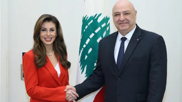 مورغان أورتاغوس (يسار) مرتدية لباساً رسمياً أحمر وخاتم نجمة داوود، تصافح الرئيس اللبناني جوزيف عون الذي يرتدي بدلة رسمية سوداء وخلفه علم لبنان