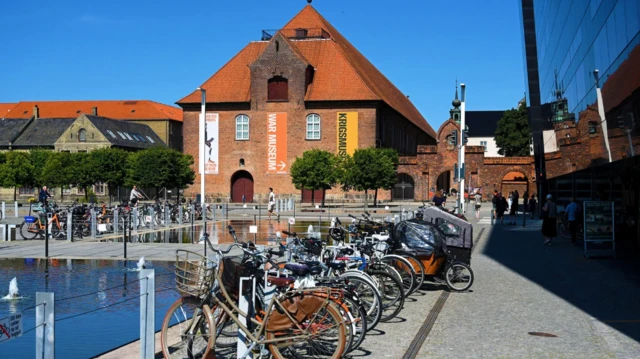 Rzędy rowerów zaparkowanych przed kopenhaskim Muzeum Wojny, mieszczącym się w zabytkowym ceglanym budynku. 